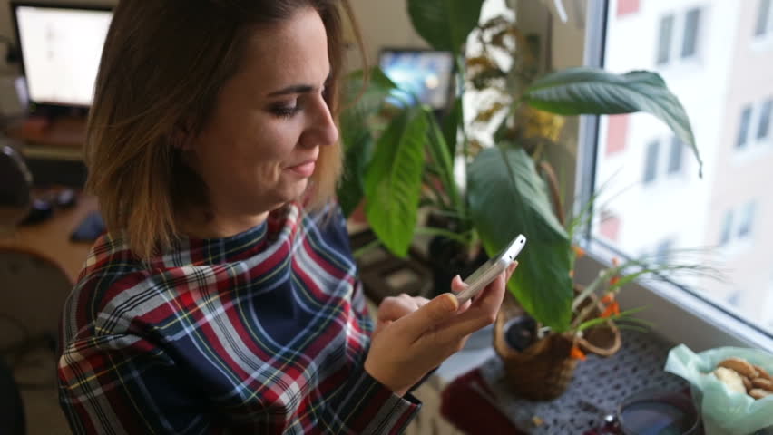 The girl is waiting for guests, she looks out the window of the house, calling on the smartphone Khu. Top-down view, closeup, sunset, natural light, indoors, real time