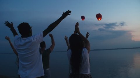 Tilt down of group of friends standing on beach at dusk and waving while watching sky lanterns fly away స్టాక్ వీడియో