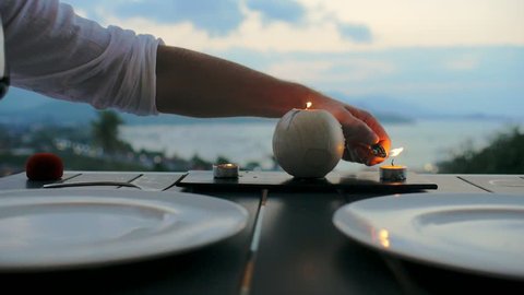 Male hand using lighter for candle lit on romantic dinner for two. Man lighting candle on dinner table on background sea landscape