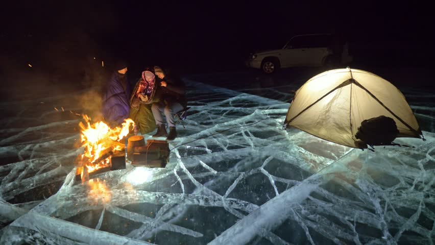 Three travelers by fire right on ice at night. Campground on ice. Tent stands next to fire. Lake Baikal. Nearby there is car. People are warming around campfire and are dressed in sleeping bags. This