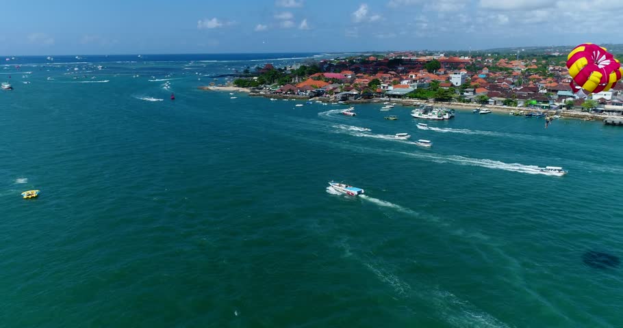 Drone footage of a speed boat towing its tandem parasail in the chanel between the south of Serangan island and the tip of Tanjung Benoa. The camera is following the boat starting behing it and passin