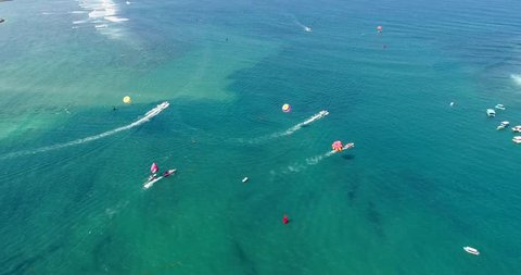 Drone footage of speed boats towing their parasail along the colorful shallow water of the south of Serangan island. The camera is facing down at 4 boats going in the same direction and is descending