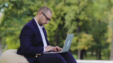 Young disheartened smart man businessman or student in casual blue shirt, glasses sitting at table, prints on a laptop in city park. Crypto investor working outdoors. Crypto-currency operation.