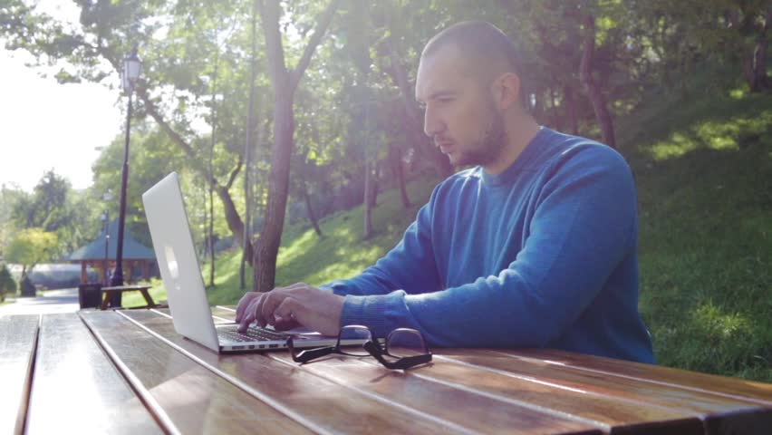 Young disheartened smart man businessman or student in casual blue shirt, glasses sitting at table, prints on a laptop in city park. Crypto investor working outdoors. Crypto-currency operation. | Shutterstock HD Video #1009552931