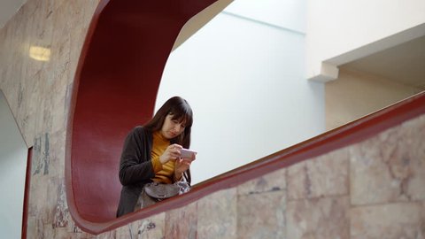 A young attractive woman sits in a niche and prints a message using a smartphone indoors with marble walls.