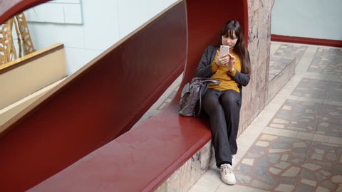 Happy attractive young woman sits in a niche and takes a selfie on a white smartphone indoors with marble walls.