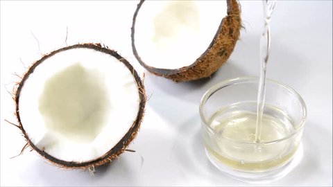 Pouring coconut oil in a cup with split coconuts on a white background.