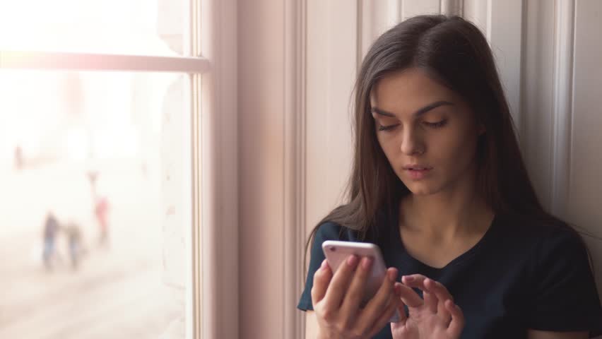 Beautiful dreaming woman typing a message near the window, using the gadget with great pleasure, inloved girl chatting on phone in cozy apartment on ordinary weekday | Shutterstock HD Video #1009589744
