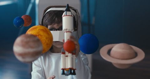 CU Cute little boy wearing cardboard astronaut helmet flying toy rocket through planets, exploring deep space. 4K UHD 60 FPS SLOW MO