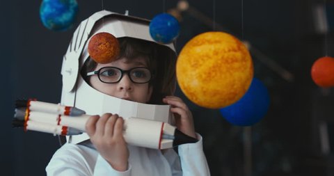 CU Cute little boy wearing cardboard astronaut helmet flying toy rocket through planets, exploring deep space. 4K UHD 60 FPS SLOW MO