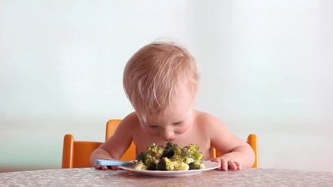 baby clap his hands and eats with pleasure steamed broccoli by fork and hand, happy kid, white background  