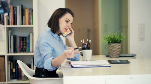 Young businesswoman with documents talking on cellphone sitting at table in home
