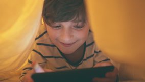 A happy smiling boy lies in bed under a blanket and plays enthusiastically on a smartphone in a game in the dark. The face of the child is lit by a bright monitor
