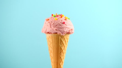 Ice cream cone close-up. Pink Icecream scoop in waffle cone rotated over blue background. Strawberry or raspberry flavor Sweet dessert decorated with colorful sprinkles, rotation closeup. 4K UHD