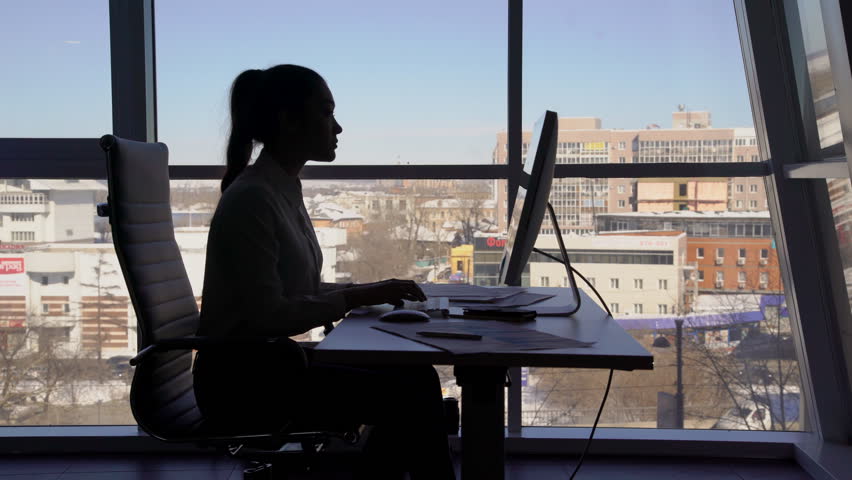 Silhouette of business woman who works at computer and talking on phone near window in office. Concept: window in office, business woman, computer and telephone