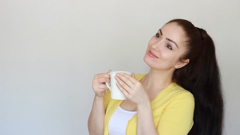Portrait of a beautiful brunette girl who drinks a hot drink from a cup, wich holds in his hand. Young woman smiles on the white light background in studio. Close-up.