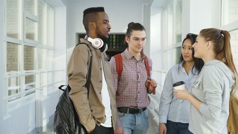 Multi-ethnic group of four students standing in wide lighty glassy hall in college talking to each other in positive way smiling ant laughing dinking coffee