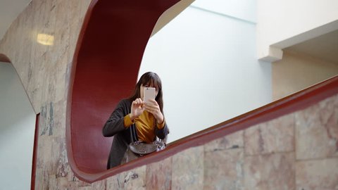 A happy attractive young woman sits in a niche and makes a video call using a white smartphone indoors with marble walls. The brunette waving her hand welcoming the interlocutor.