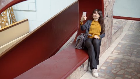 Happy attractive young woman sits in a niche and takes a selfie on a white smartphone indoors with marble walls.