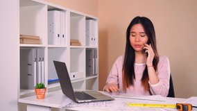 asian architect working in casual office. attractive brunette with long hair has phone conversation with client or partner checking building plan