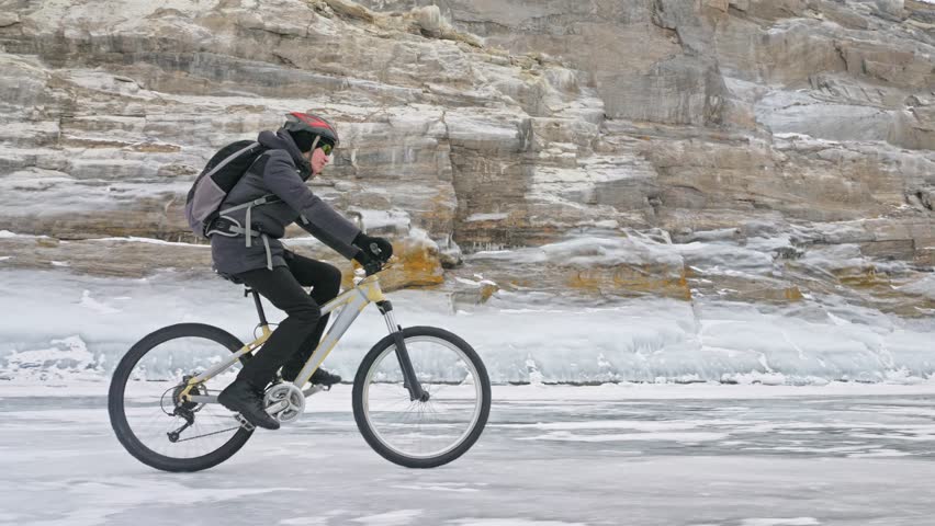 Man is riding bicycle near the ice grotto. The rock with ice caves and icicles is very beautiful. The cyclist is dressed in gray down jacket, cycling backpack and helmet. The tires on covered with Royalty-Free Stock Footage #1009671170