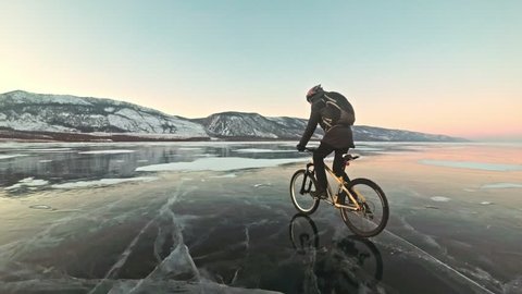Man is riding a bicycle on ice. The cyclist is dressed in a gray down jacket, backpack and helmet. Ice of the frozen Lake Baikal. The tires on the bicycle are covered with special spikes. The traveler – Stockvideo