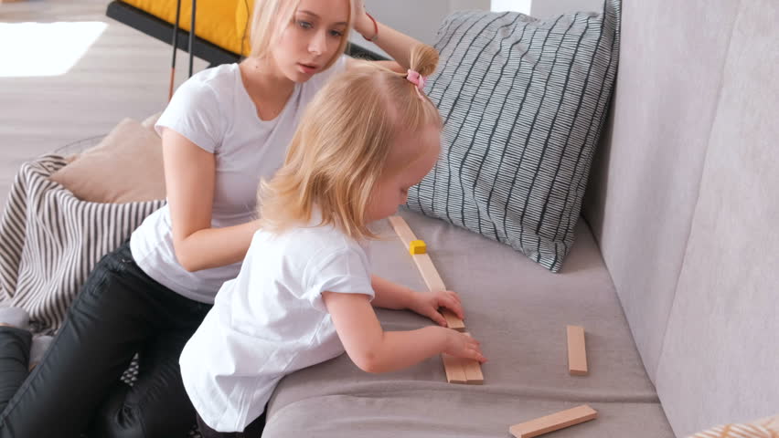 Little charming blond girl playing wood blocks with her mom on the sofa. Road from wood blocks.