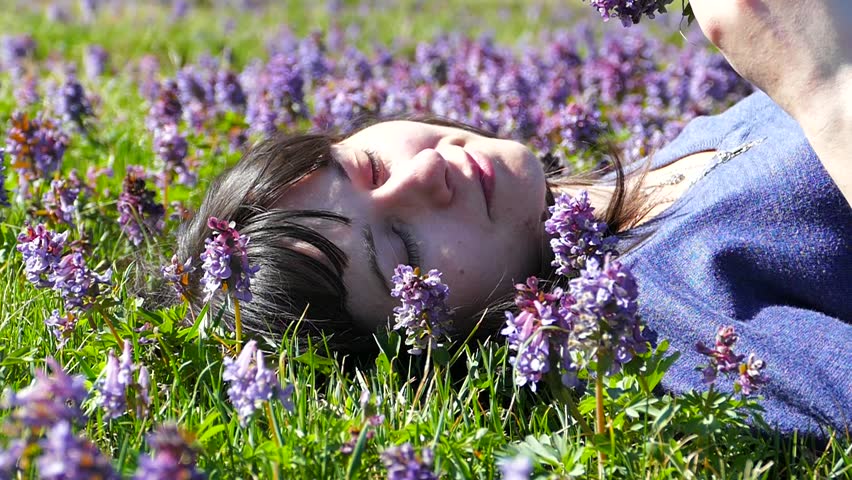 Beautiful girl relaxes on lawn lying on grass with blue flowers. A girl is resting and smiling on a spring sunny day. Hair is heard by a slight breeze.