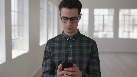 portrait of attractive young hipster man texting browsing social media using smartphone mobile technology wearing glasses in new apartment room