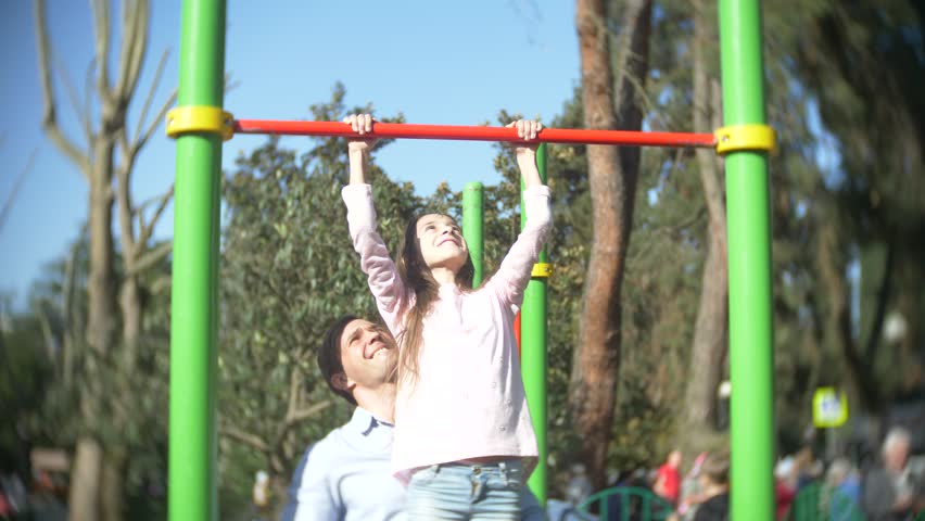 A handsome man teaches a girl in a pink T-shirt how to pull himself up correctly on a horizontal bar in a summer sunny park, 4k, slow motion