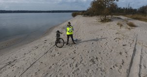 Operator of a mature man with a beard sits a drone on sand and a bicycle. Man with a beard stands on the sand with a control panel for a drone and a bicycle