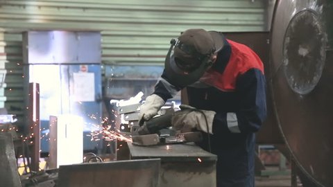 A worker in a face mask and gloves is working the metal with an angle grinder. Industrial scenery, metal processing plant, industrial tool.