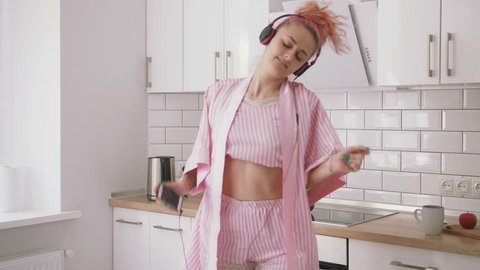 Happy young woman with pink hair dancing in kitchen wearing pink pajamas and listening to music with headphones