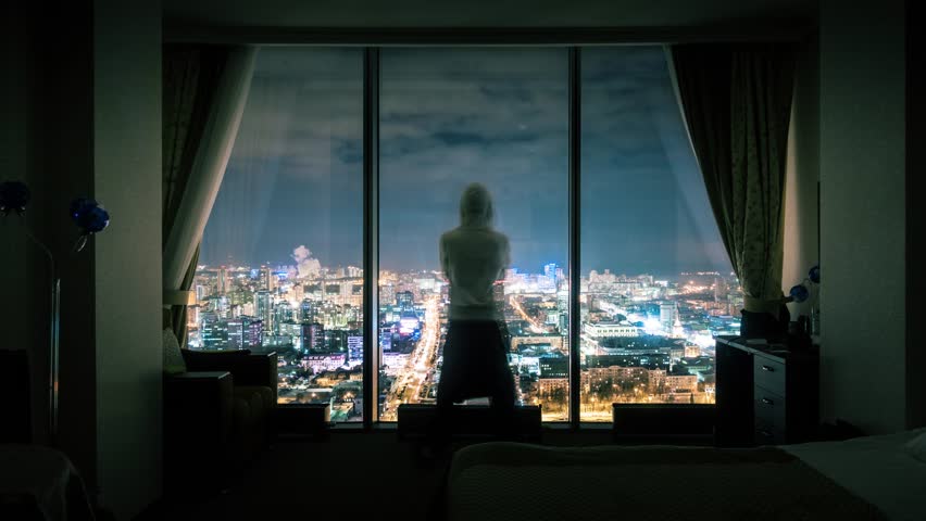 Hooded anonymous man silhouette standing in dark room looking out of panoramic window with illuminated night city view in background. Timelapse, 4K UHD. Zoom in. Royalty-Free Stock Footage #1009716761