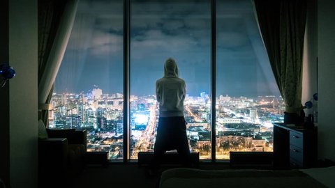Hooded anonymous man silhouette standing in dark room looking out of panoramic window with illuminated night city view in background. Timelapse, 4K UHD. Zoom in.