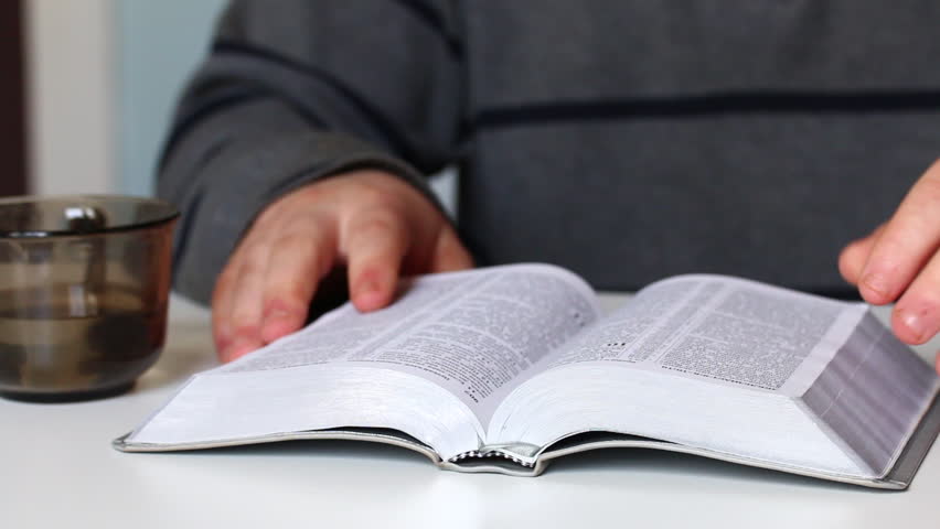 A young man reads the Bible sitting at the table. Flips through the pages.
