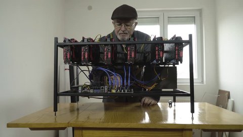 Mature caucasian male person in work coat checking cables and port connection on mining machine motherboard, graphics card in the foreground, camera crane motion to close up, concept cryptocurrency.