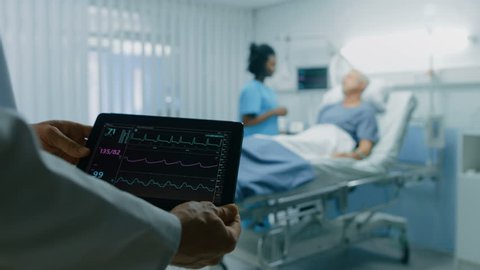 In the Hospital Doctor Holds Tablet Computer Showing Vital Signs of a Senior Patient Lying in Bed. Nurse Does Checkup. Modern Geriatrics Ward. Shot on RED EPIC-W 8K Helium Cinema Camera.