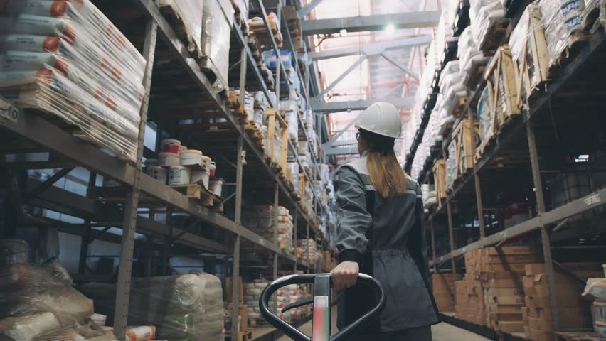 Following shot of female industrial worker in hard hat walking through along shelves with trolley for the warehouse. Female laborer in uniform passing by, pulling trolley stuffed with piled boxes | Shutterstock HD Video #1009730489