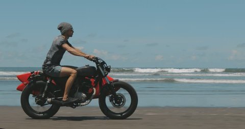 Handsome man biker surfer driving his black motorbike cafe racer with red surfboard shortboard on the beach along the ocean at sunny day. 4k video shooting by handheld gimbal