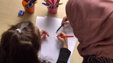 Young beautiful mother in hijab, small daughter paints with colored pencils, paints, children's coloring, family idyll concept, close up, top shot 50 fps