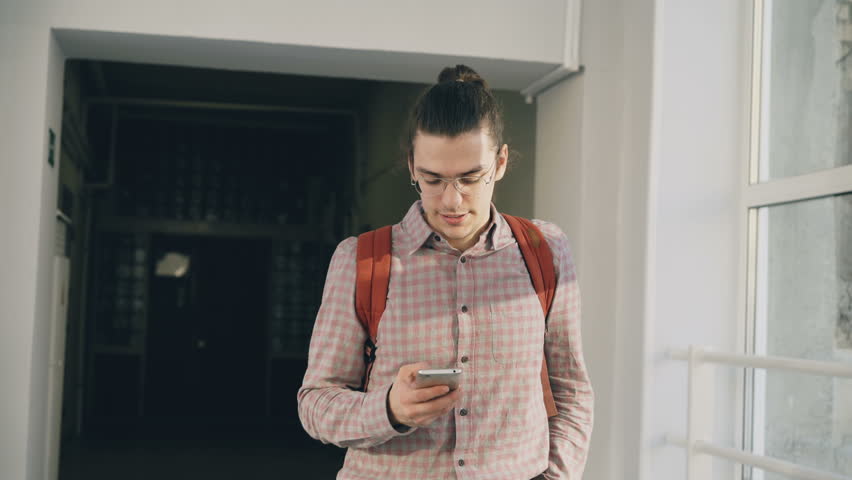 Young handsome caucasian hipster male student is walking in white lighty spacious corridor in college holding phone making phone call passing by other students