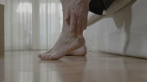 Patient mature man with arthritis sitting on bed and rubbing his ankle and foot relieving the ache
