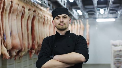 Portrait of handsome young smiling butcher in black uniform at the meat manufacturing with pork carcasses and brighgt light on background factory male worker crossing arms confident man butchering