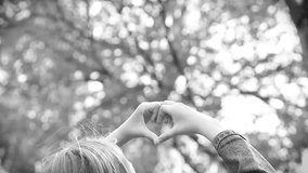 Back view of young pretty teen girl forming heart with her hands against blurry green fresh foliage bokeh of spring trees outdoors. Love and nature concept. Black and white video footage.