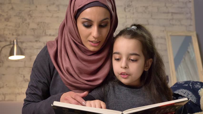 Young woman in hijab sits on sofa with her daughter and teaches her how to read, book, happy family concept, close up, home comfort in the background 50 fps
