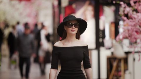 A young elegant rich girl in black dress and hat walks along the pedestrian shopping Mall street past the show rooms and boutiques. Slow motion