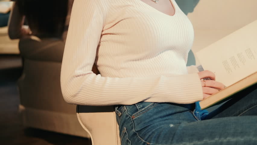 Young pretty woman with long hair sits on a chair and reading a book in the apartment. Slow motion