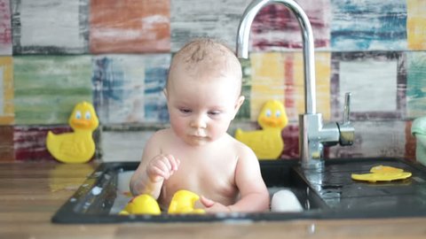Cute smiling baby taking bath in kitchen sink. Child playing with foam and soap bubbles in sunny kitchen with rubber ducks, little boy bathing, fun with water.