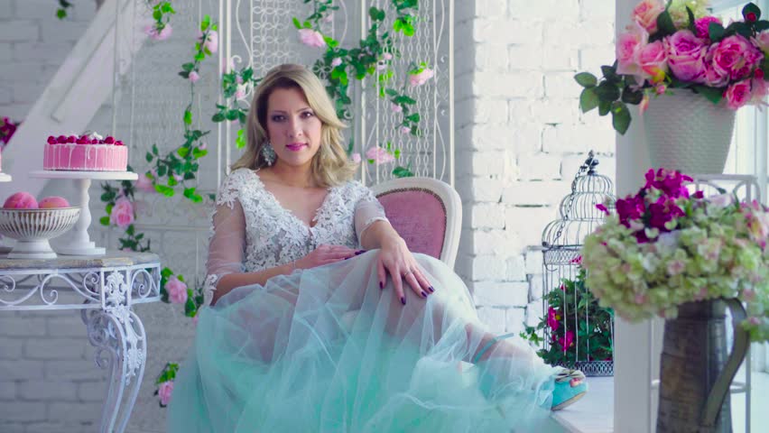 Beautiful model in peignoir sitting in a chair against white brick wall with flowers.